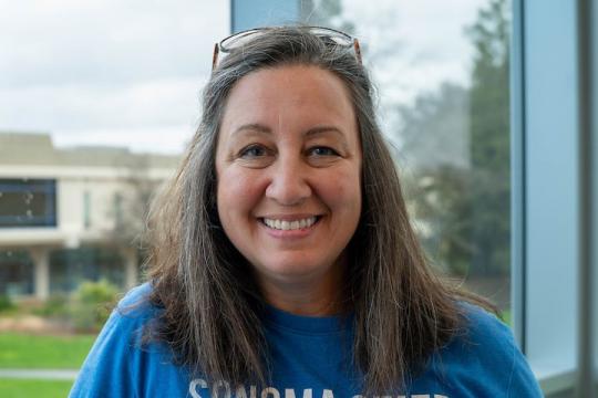 Elizabeth stands in front of a window, smiling at the camera. She wears a blue shirt, her shoulder-lengeth hair is down but is tucker behind her ears by her glasses which sit atop her head. 