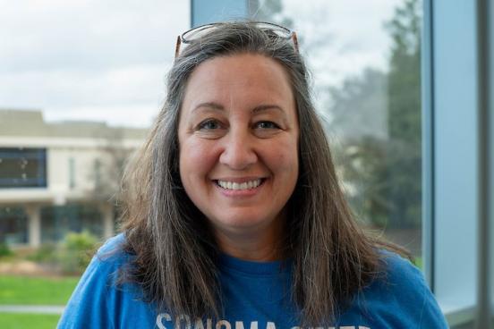 Elizabeth stands in front of a window, smiling at the camera. She wears a blue shirt, her shoulder-lengeth hair is down but is tucker behind her ears by her glasses which sit atop her head. 