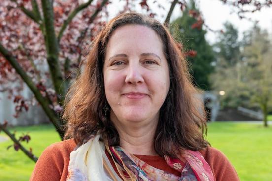 Gillian stands outside, with a tree with spring blooms and bring green grass behind her. She looks directly at the camera with a small smile. Her hair is down and she wears an orangey-red sweater and multi-colored light scarf. 
