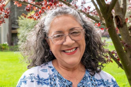 Giselle stands outside, green grass and a bloom tree are visible behind her. She smiles directly at the camera, her head slightly tilted. She wears glasses and a blue and white patterned shirt