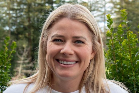 Lauren smiles brightly directly at the camera. She wears a white shirt and her hair is down, tucked behind her ears. Behind her is a line of redwood trees. 