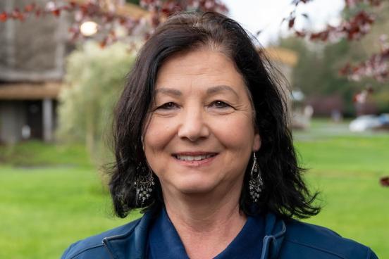 Lucia stands outside, giving the camera a small smile. Her dark, shoulder length hair is down and she wears a blue collared shirt. Behind her you can see green grass and blooming trees.