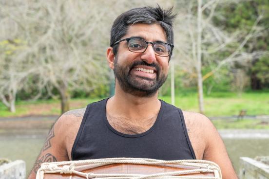 Raghav stands outside, a lake and blooming trees behind him. He wears glasses, a tank top, and is holding a small bongo drum. He's looking directly at the camera and is giving a big smile. 