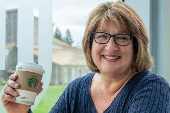 A close photo of Yvonne as she sits at a table in front of windows in the Student Center. She smiles at the camera warmly while holding a coffee cup in her left hand. 