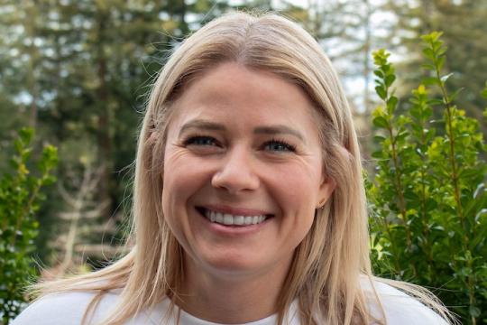 Lauren smiles brightly directly at the camera. She wears a white shirt and her hair is down, tucked behind her ears. Behind her is a line of redwood trees.