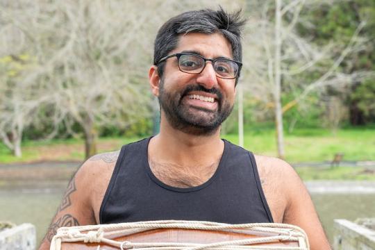 Raghav stands outside, a lake and blooming trees behind him. He wears glasses, a tank top, and is holding a small bongo drum. He's looking directly at the camera and is giving a big smile.